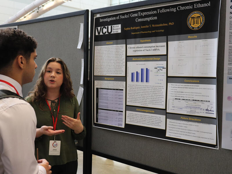 A photo of a woman and man standing to the right of a research poster. The woman is speaking and the man is listening to her. 