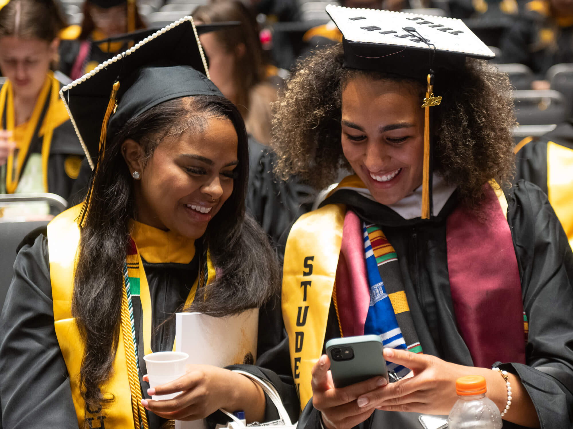 Two VCU graduates in graduation robes smile while looking at a phone.