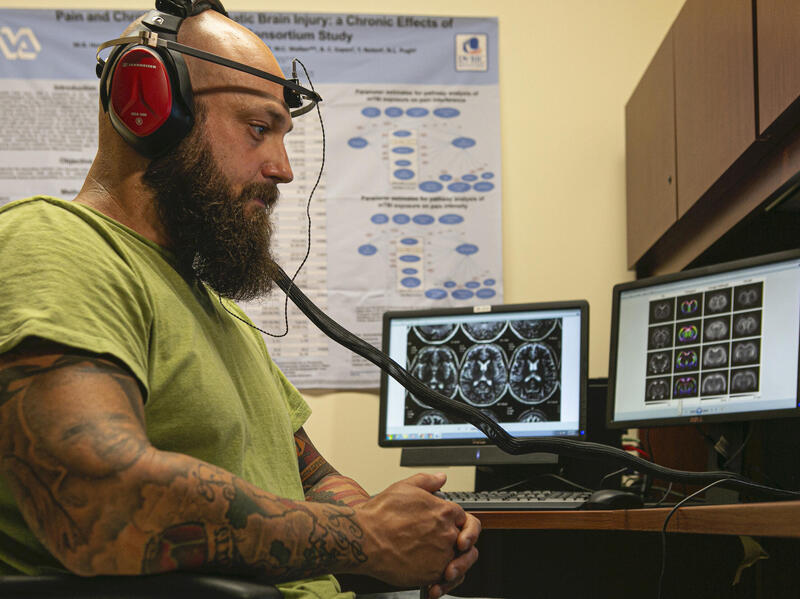 A person wearing headphones and a band around their head sits in a chair. Computer screens display images of brain scans.