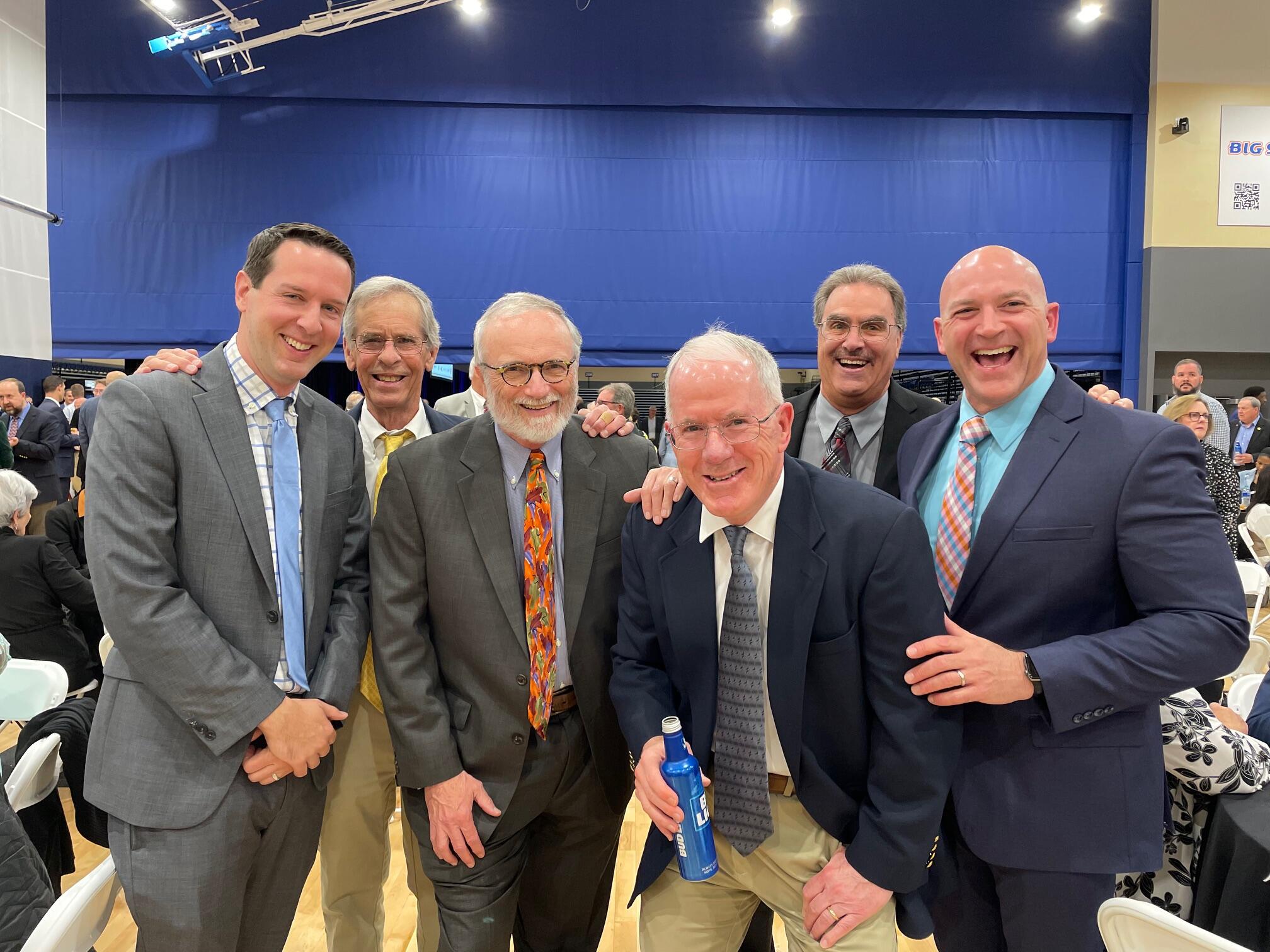 A group photo of six men wearing suits and ties standing in a room full of people 