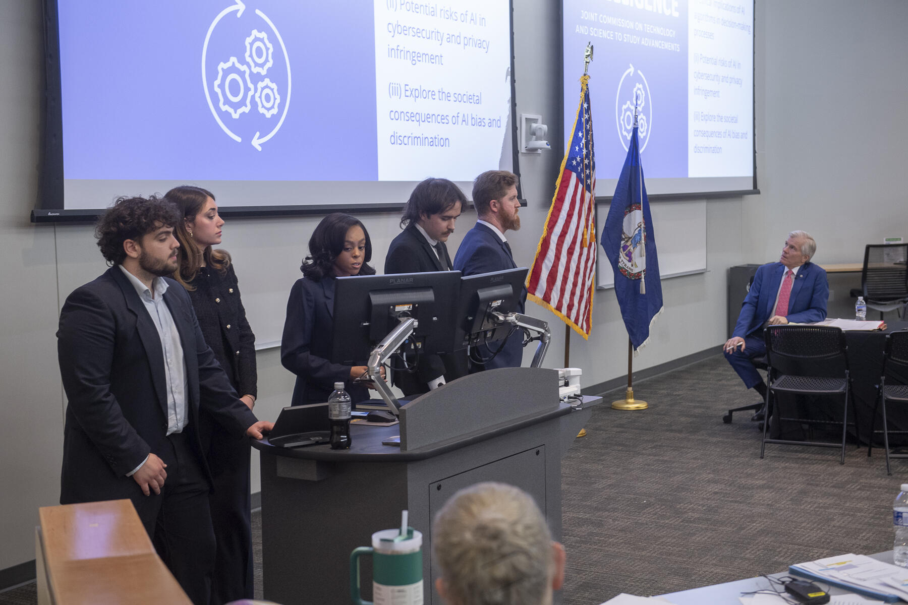 A photo of five people standing behind a podium and in front of two screens with projections on them. To the right of the group is an American flag and a Virginia state flag, along with a man sitting in a chair looking up at the screens.