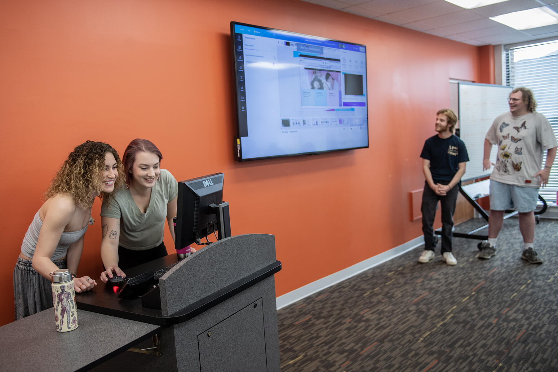 Four people standing at the front of a room giving a presentation. On the left two people are looking at a computer and on the right two other people stand up facing a large TV screen. 