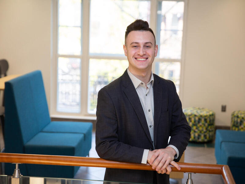 A man wearing a suit smiling at the camera. His hands are resting on a wooden banister. 