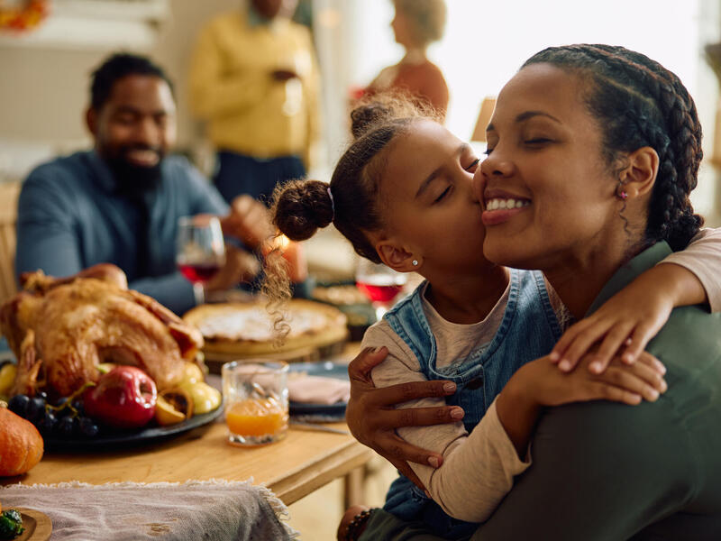 A photo of a family at the Thanksgiving dinner table. A little girl is kissing her mom on the side of her face. A dad is sitting across from them, smiling. 