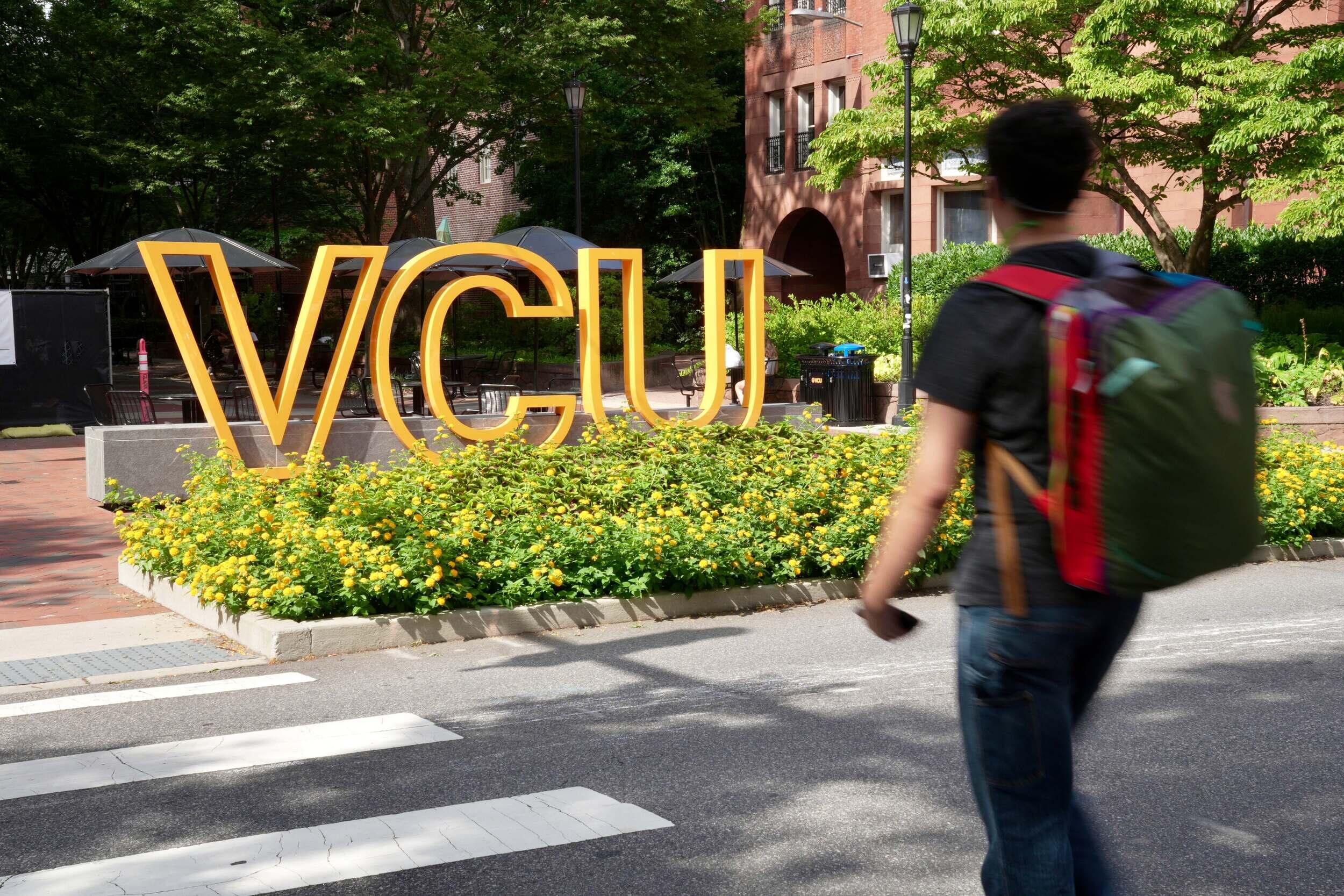 A photo of a person wearing a backpack crossing the street at a crosswalk. On the other side of the street is a sign that says \"VCU\" in large yellow letters. 