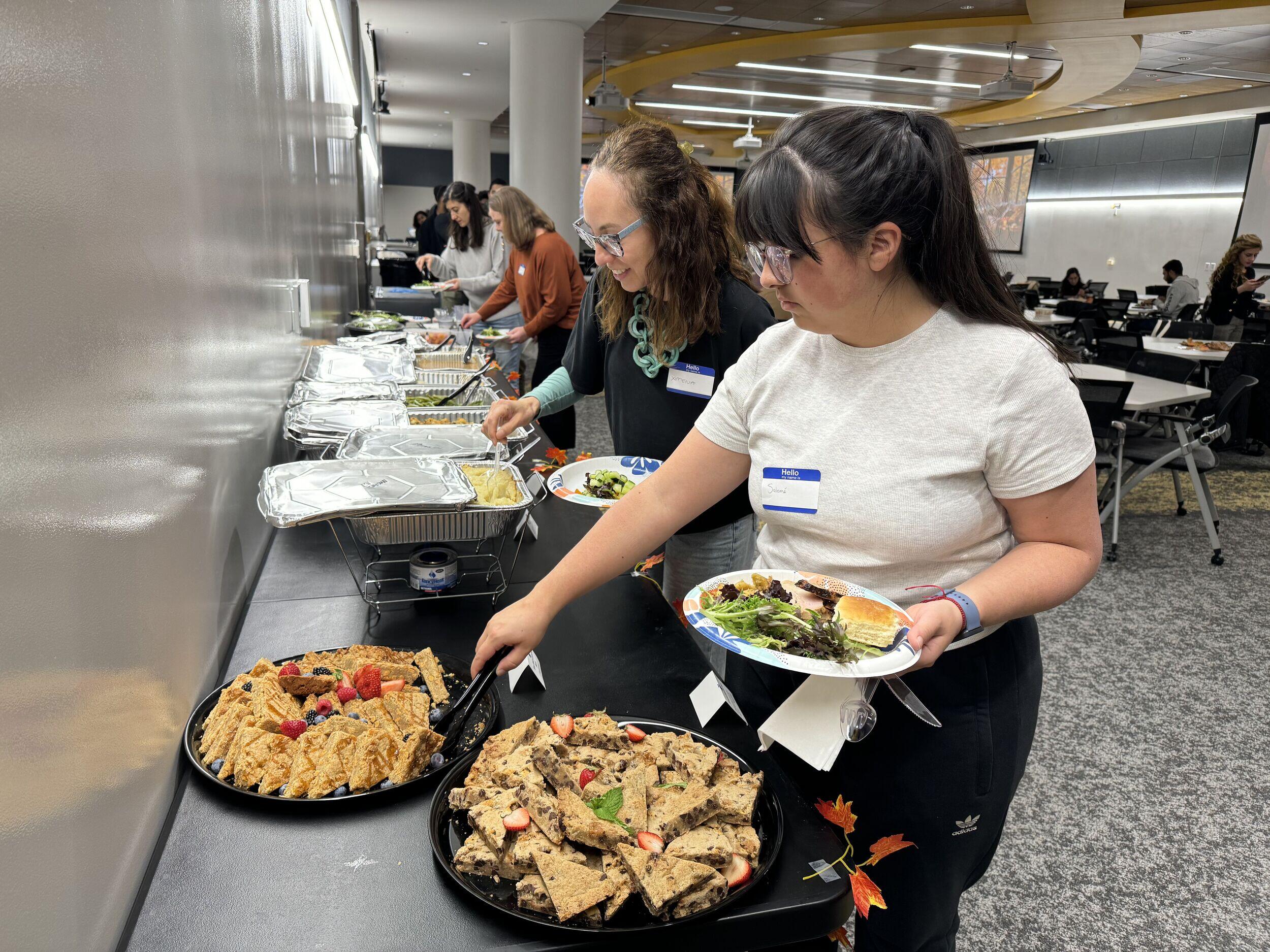 A photo of a row of tables against a wall with platters of food on them. There are four people along the line of tables who are taking food off of the platters. 