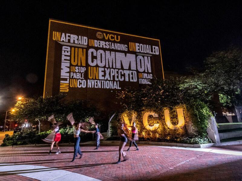 People walking across a plaza at night with lit up letters behind them that spell \"VCU.\" Above the letters is a large banner that says \"UNCOMMON\" in yellow and black text.