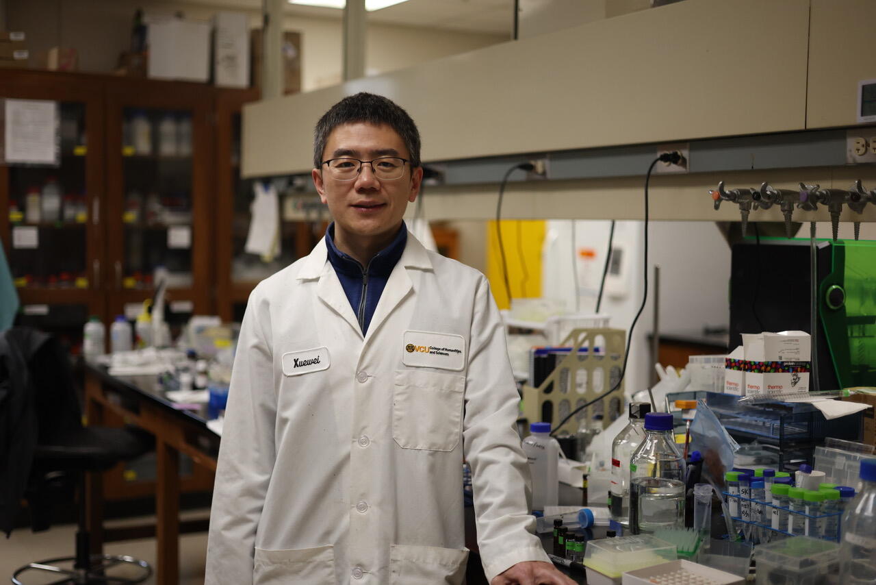 A photo of a man wearing a white lab coat standing in a labrartory. 