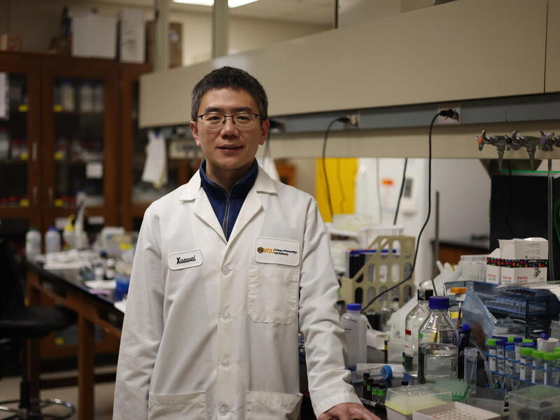 A photo of a man wearing a white lab coat standing in a labrartory. 