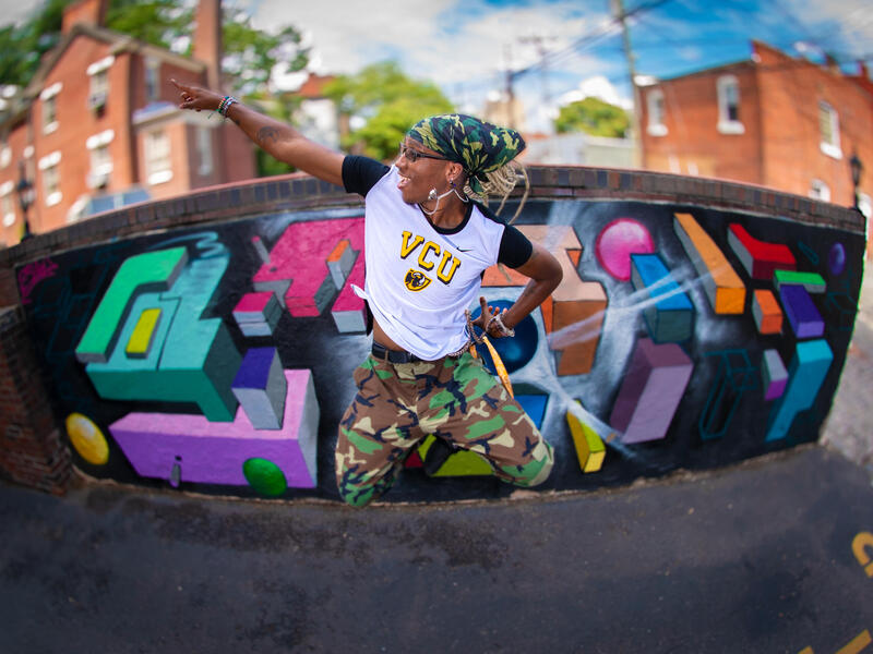 A woman in a VCU shirt and camouflage pants jumps in front of a colorful mural and points upward.