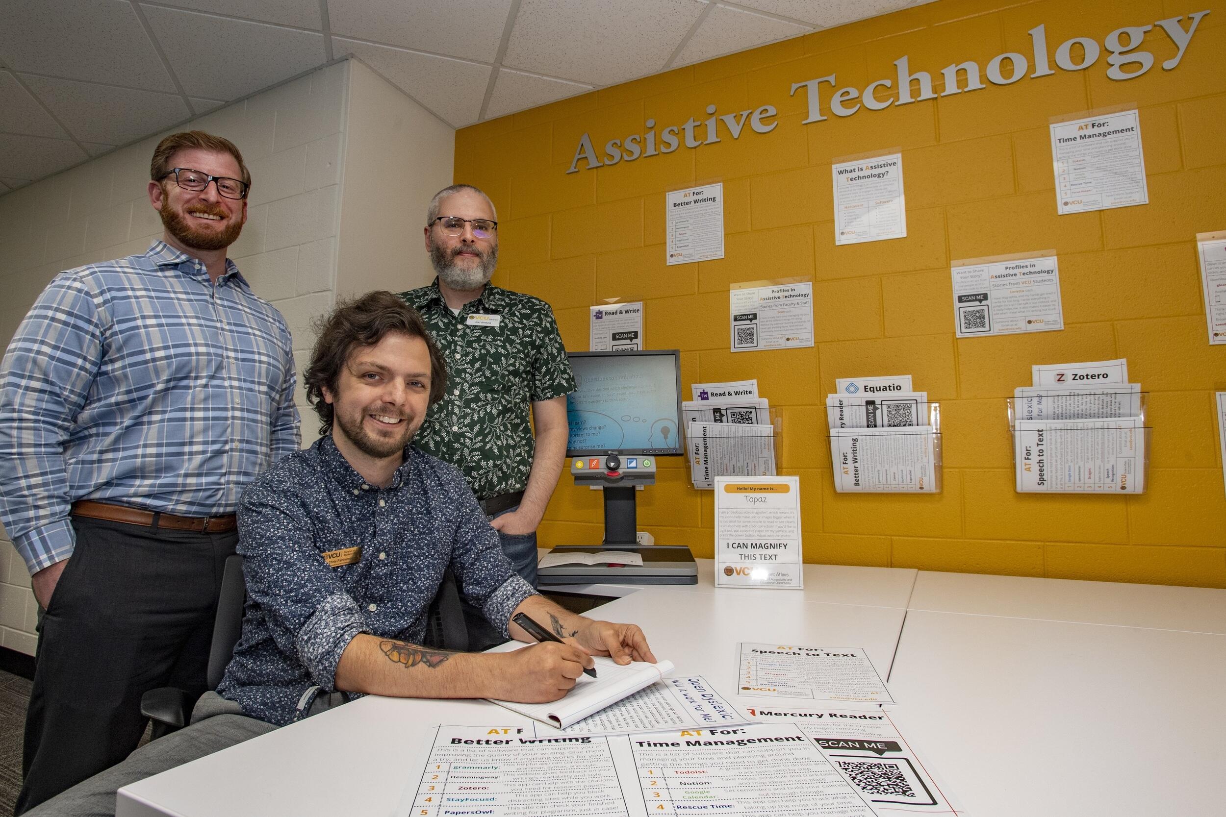 One man sitting at a table and two other men are standing behind him. They are in front of a yellow wall that says \"Assistive Technology.\"