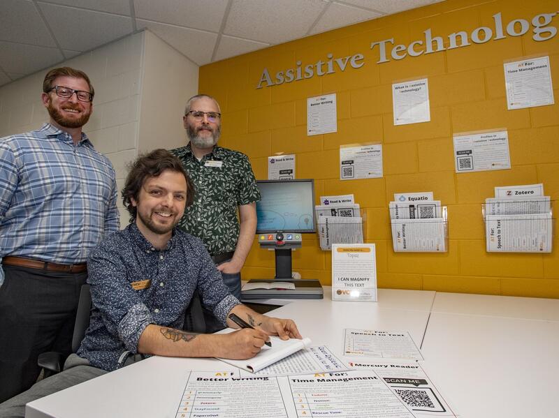 One man sitting at a table and two other men are standing behind him. They are in front of a yellow wall that says \"Assistive Technology.\"