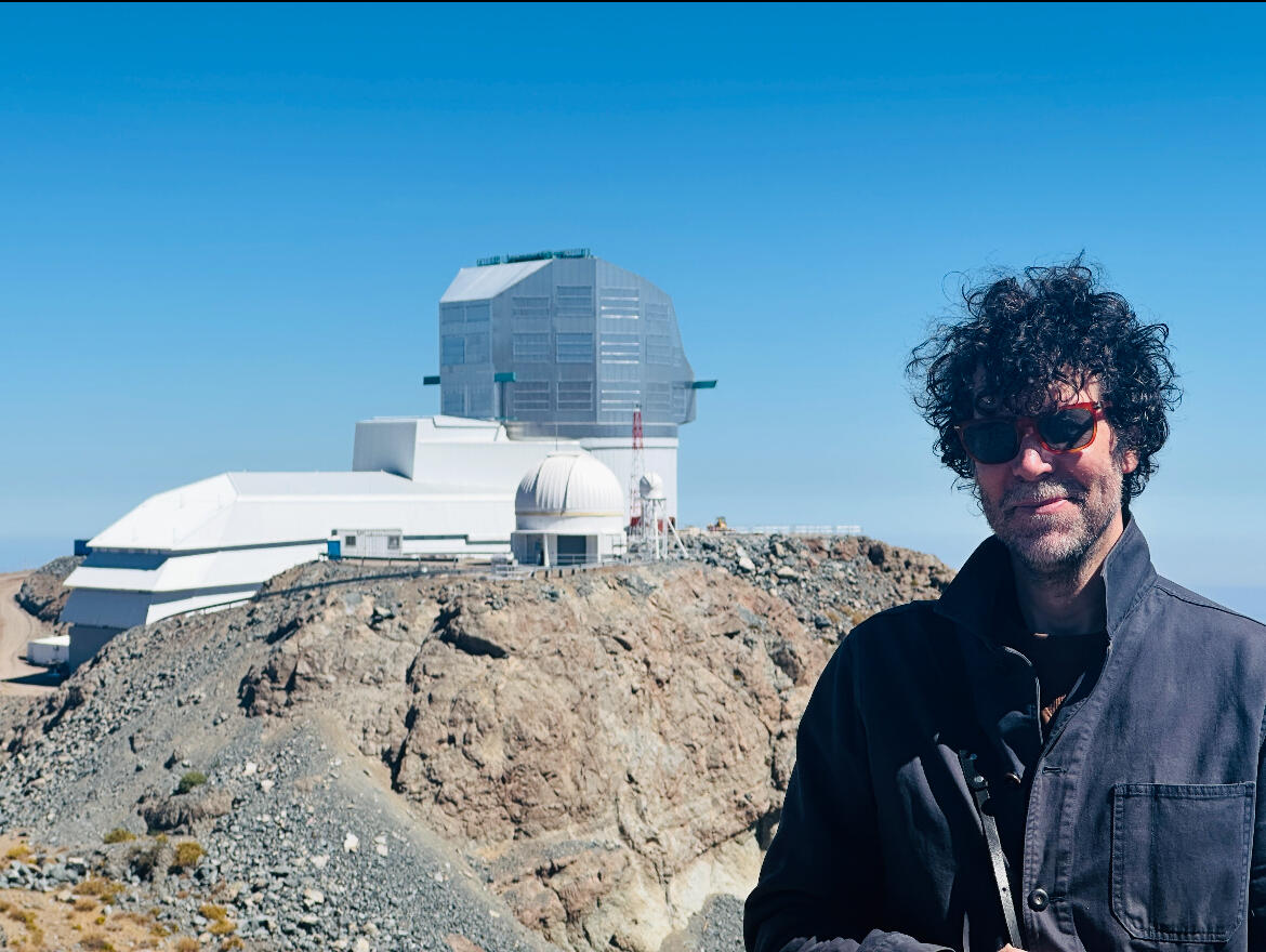 A photo of a man from the chest up. Behind him is an observatory sitting on a rocky mountain top. 