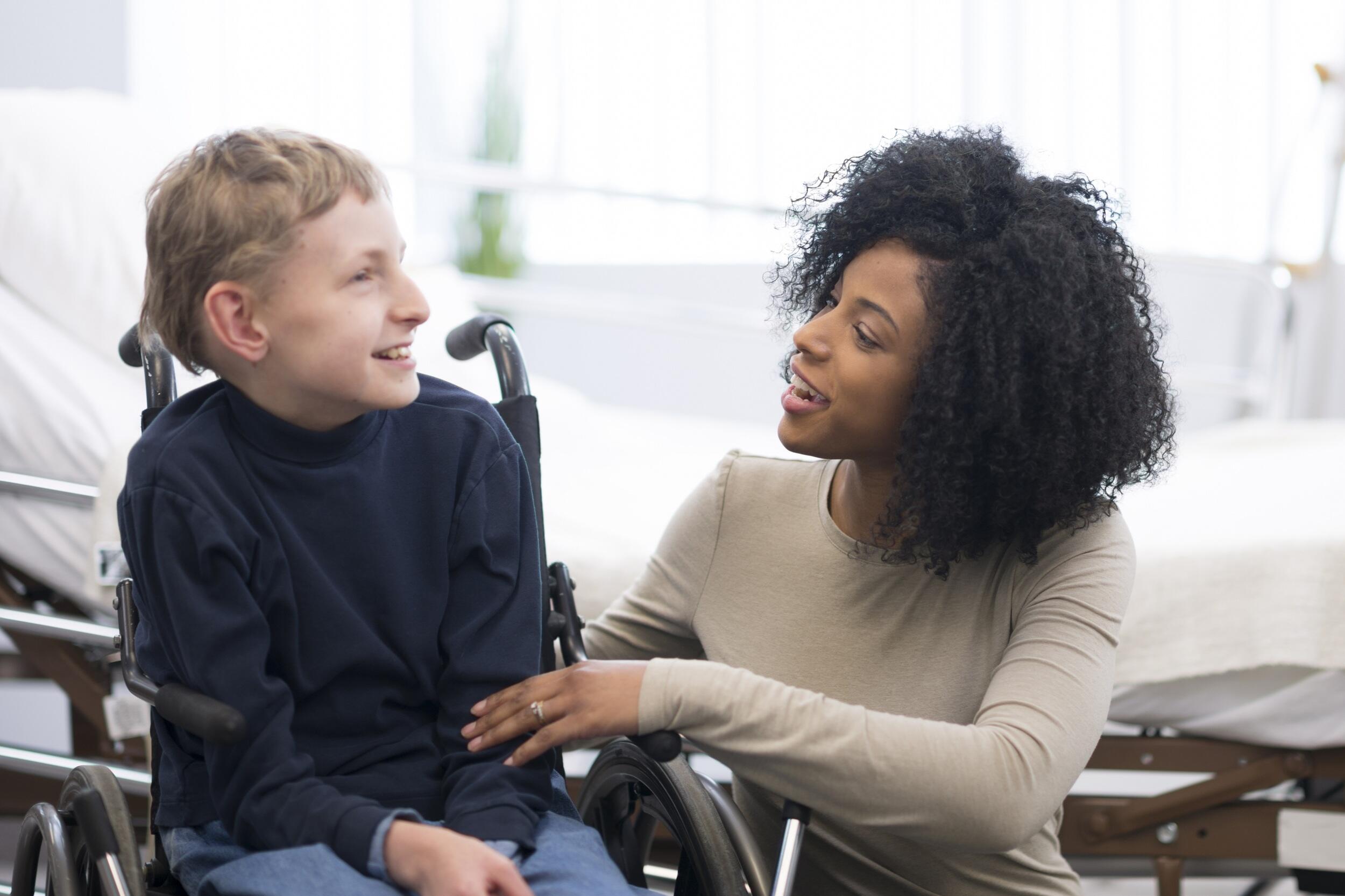 A child in a wheel chair sitting to the left of a woman kneeling down next to him. 