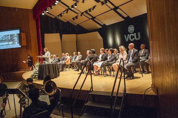 A faculty member speaks at a podium on an auditorium stage. Faculty members and administrators are seated behind her on the stage.