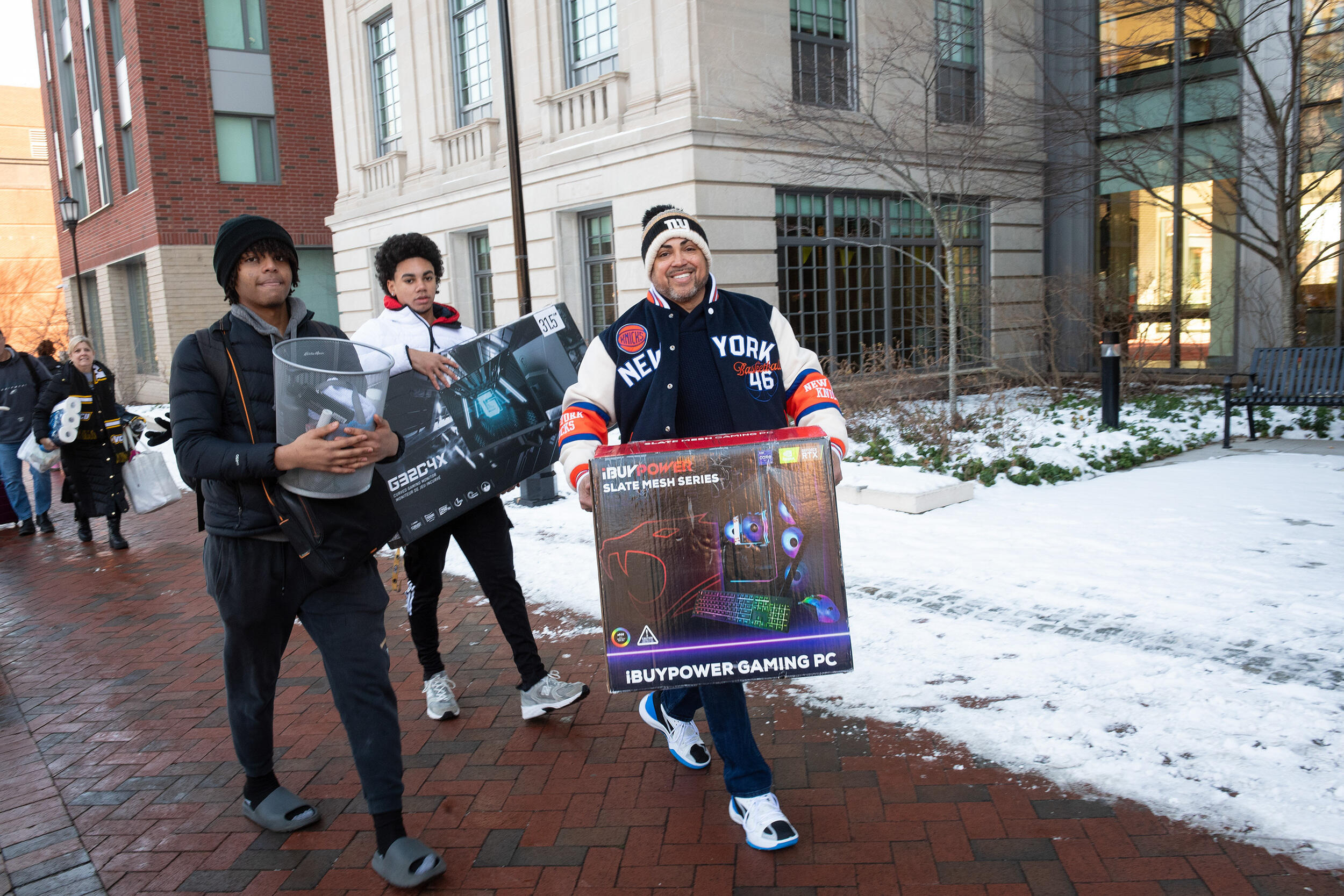 A photo of three men walking on a sidewalk. Two are carrying boxes and one is carrying a small metal trash can. 