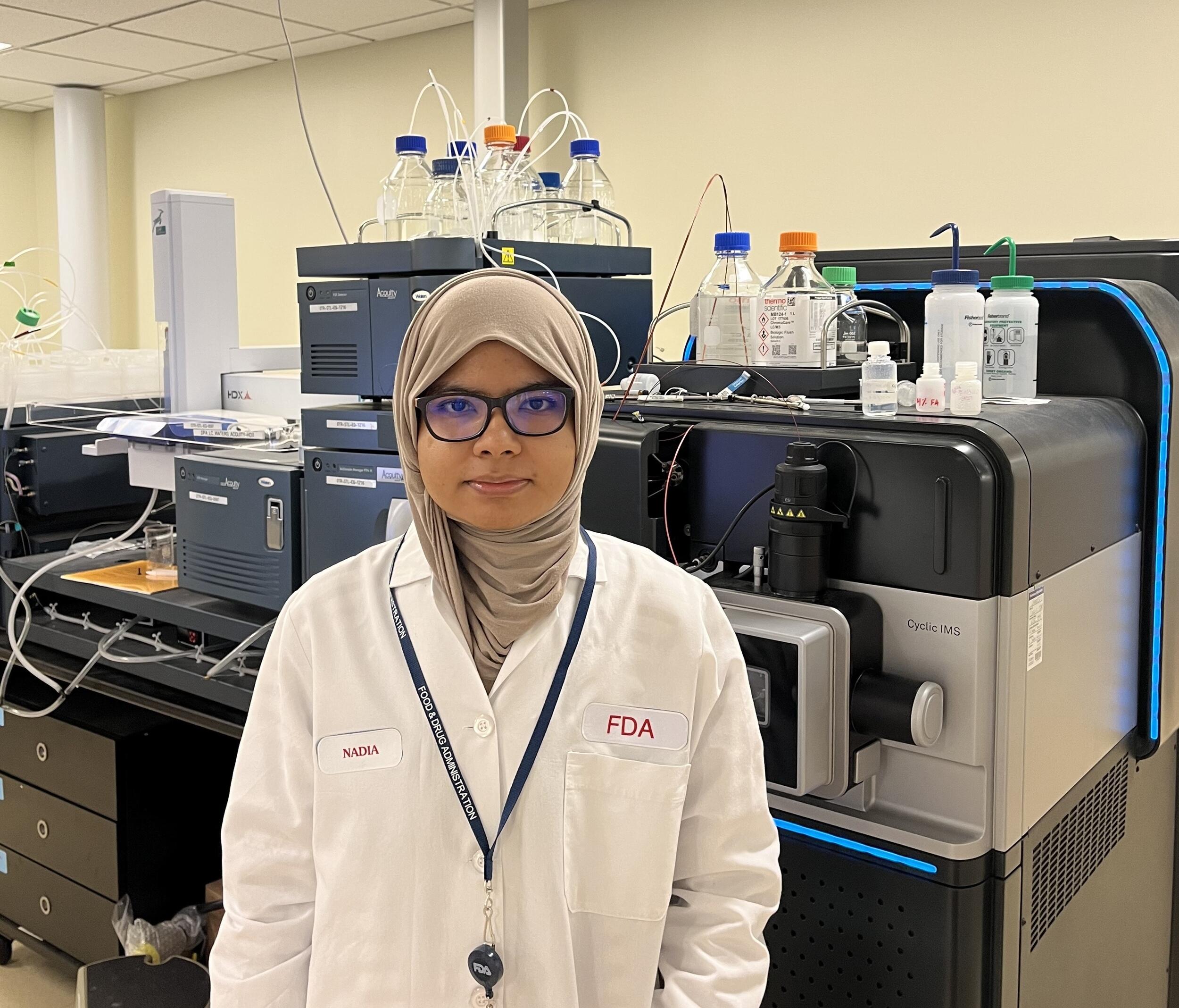 A photo of a woman from the waist up. She is weraing a white lab coat, a hijab, and glasses. 