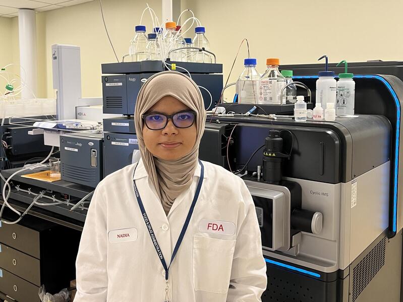 A photo of a woman from the waist up. She is weraing a white lab coat, a hijab, and glasses. 