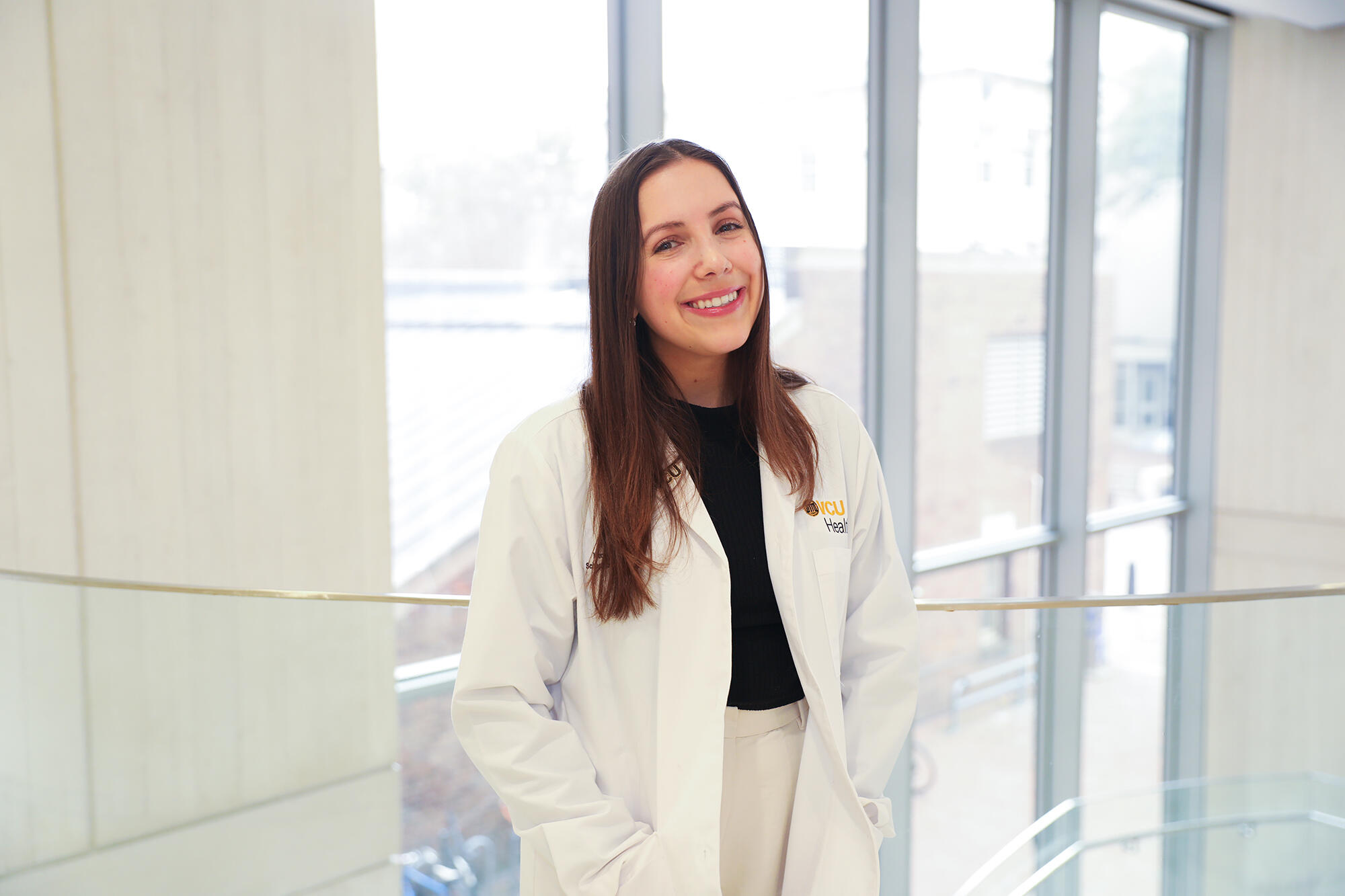 A photo of a woman standing and smiling. She is wearing a white medical coat and her hands are in her pockets. 
