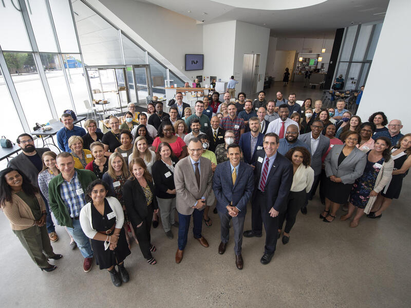A group of people standing in an open room next to windows 