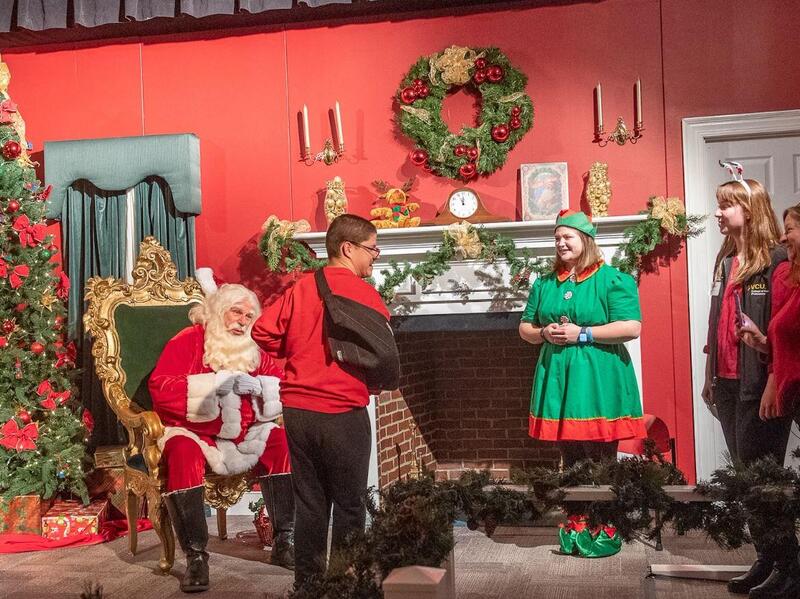 A santa sitting in a chair with a child wearing an arm brace standing in front of him. An elf and two women stand to the right of them. 