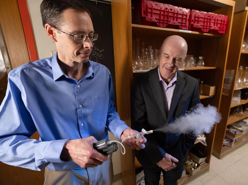 A photo of two men standin in front of book cases. The man on the right is holding a small hand held machine that is blowing a white cloud out of a tube.