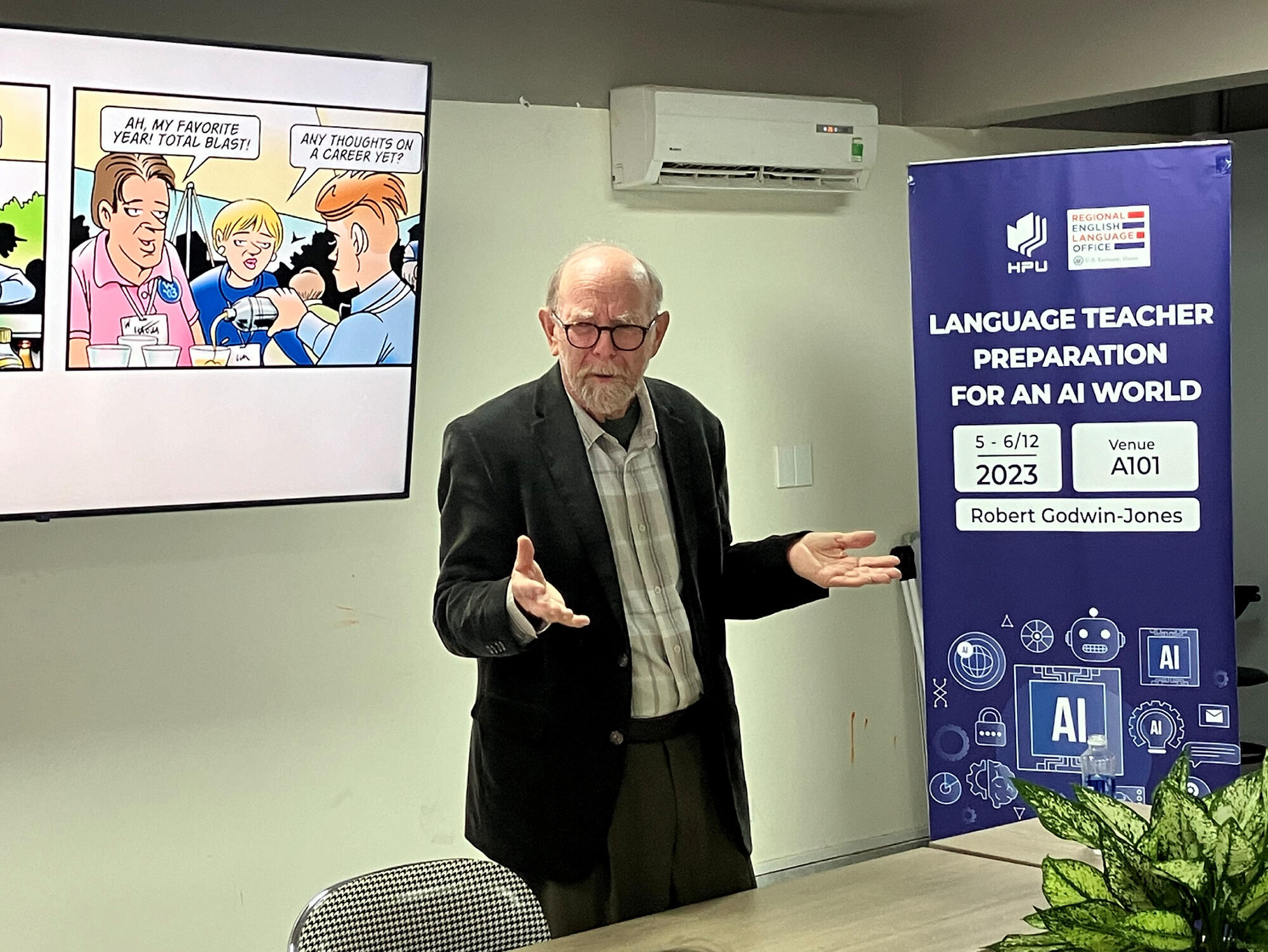 A photo of a man standing behind a table talking. Next to him is a banner that says \"LANGUAGE TEACHER PREPARATION FOR AN AI WORLD\" 