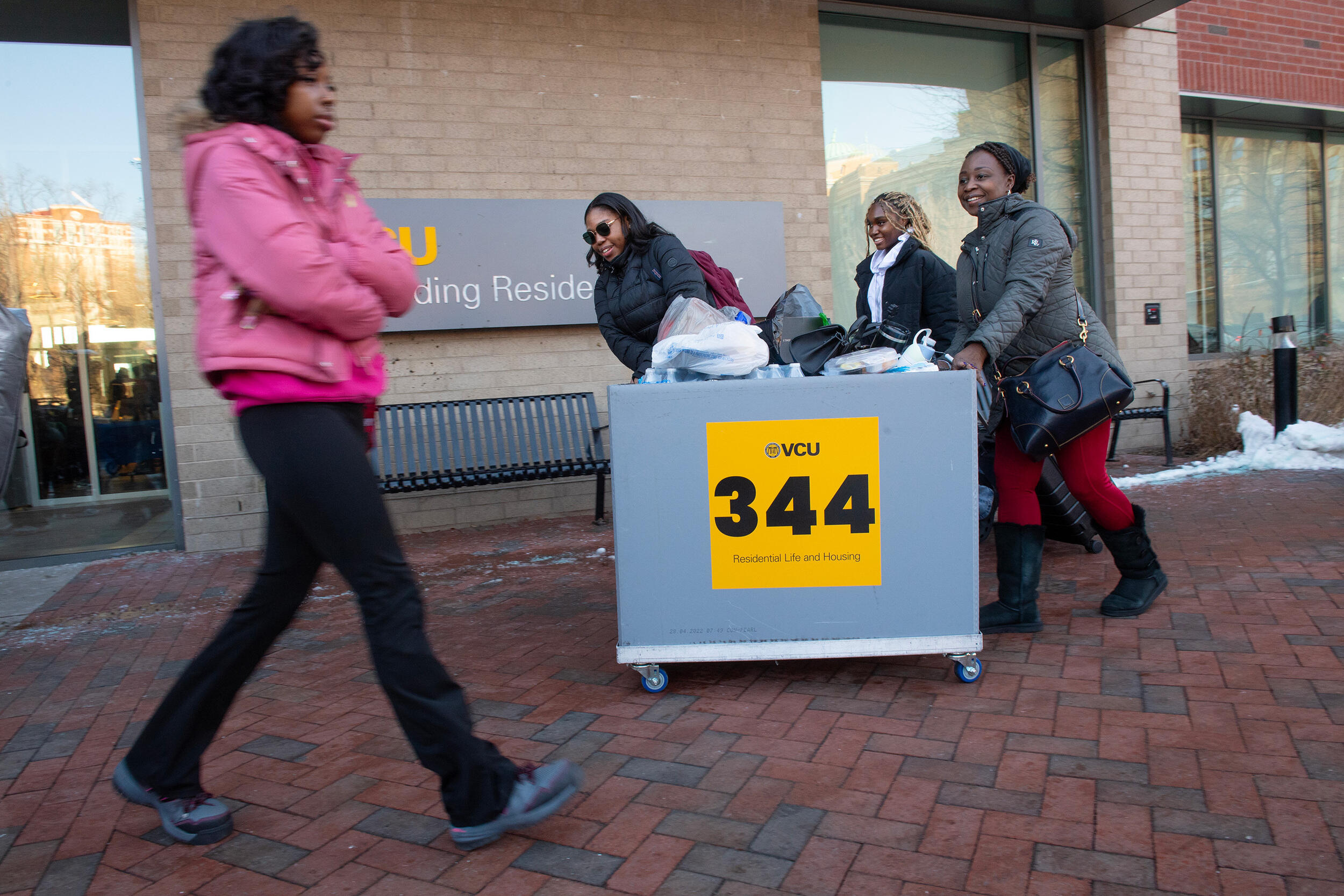 A photo of a three woman walking to the left with a rolling bin and another woman walking to the right with her arms crossed against her chest. 
