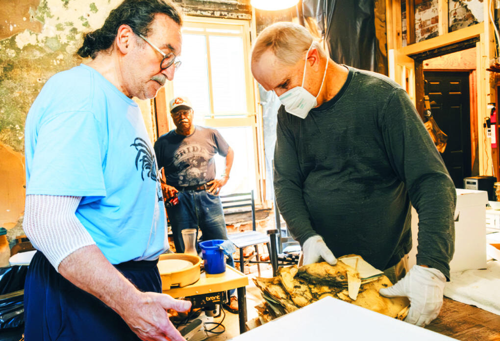 A photo of three men standing in an old room. The man on the right is wearing a face mask and latex gloves while holding a stack off old and damaged papers. 