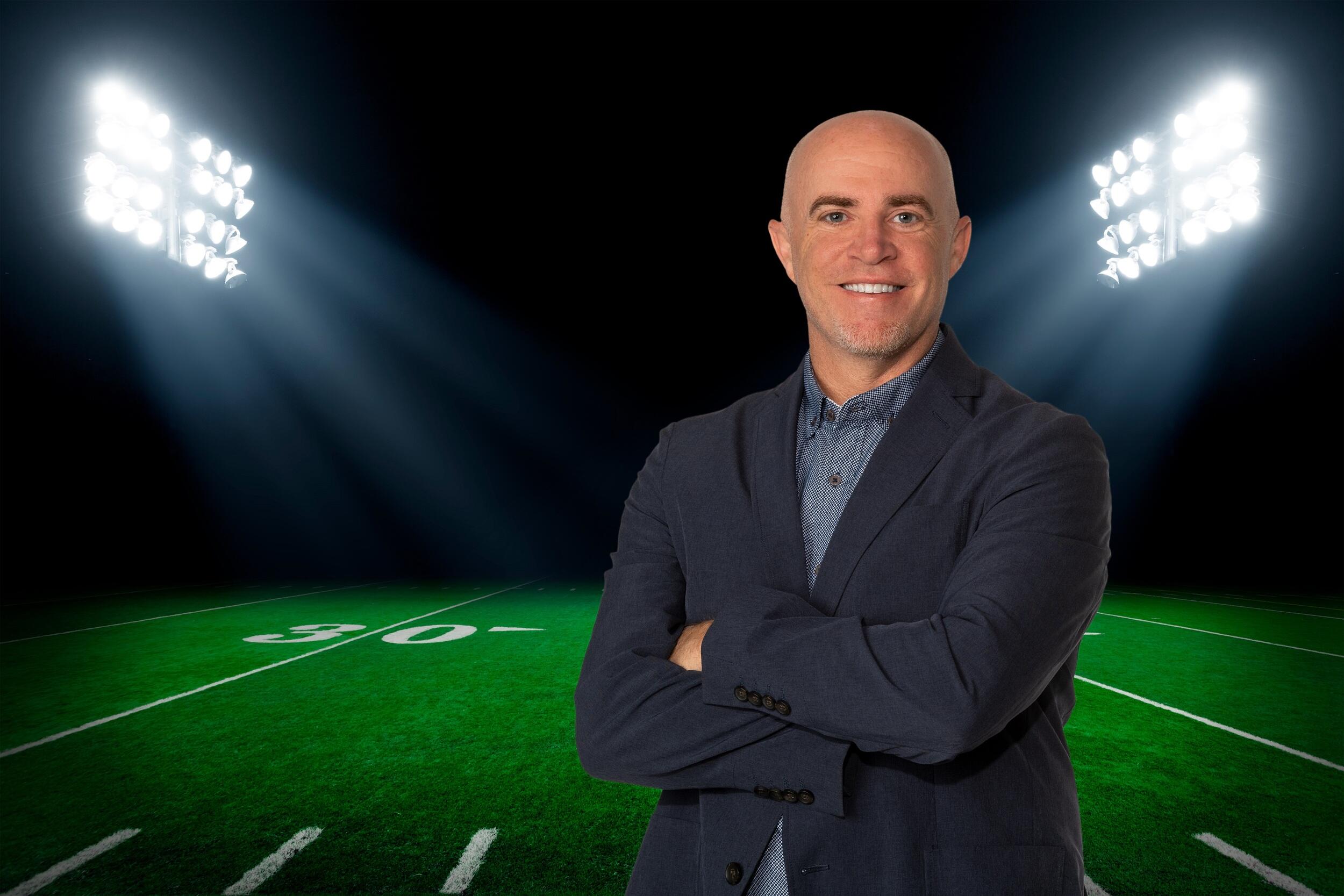 A photo of a man with his arms crossed standing on a football feild. The space behind the feild is black and two large sets of lights are illuminating the feild. 