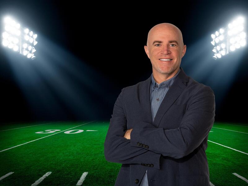 A photo of a man with his arms crossed standing on a football feild. The space behind the feild is black and two large sets of lights are illuminating the feild. 