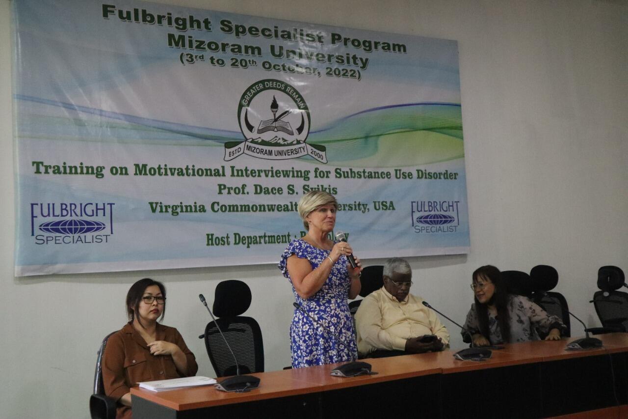 A woman standing up at a long panelist table and speaking. A woman sits to her left, and a man and women sit to her right. 