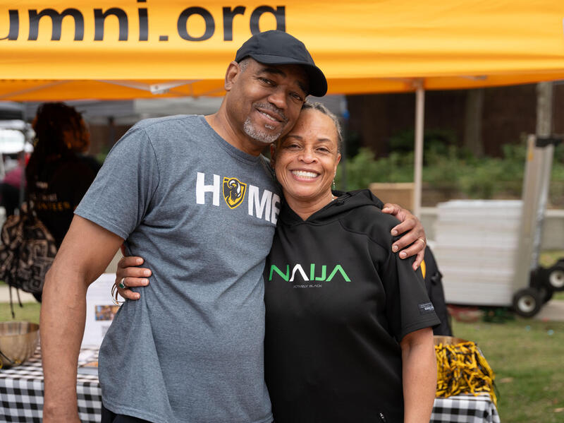 A photo of a man and woman standing next to each other. The man has his arm around the woman's shoulder, and the woman has her arm around his torso. 