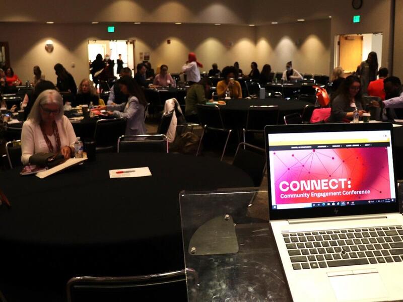 A photo of a laptop sitting on a podium in a room full of people. The screen of the computer says \"CONNECT.\" in white letters