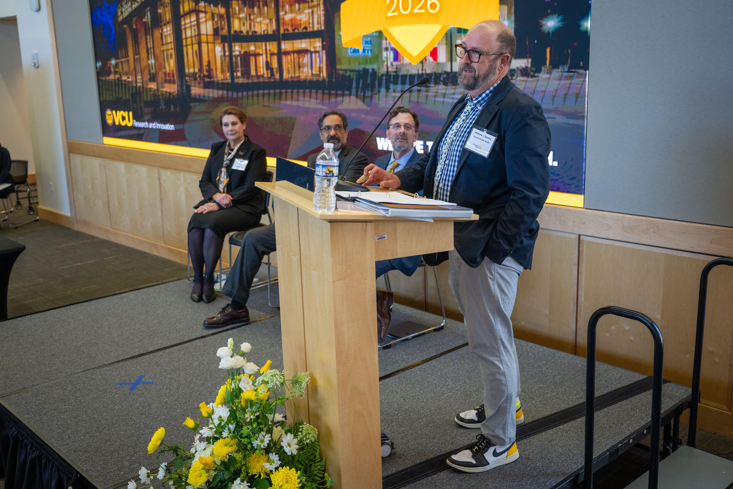 A photo of a man standing on a stage behind a podium. Next to him are three people sitting in chairs. 