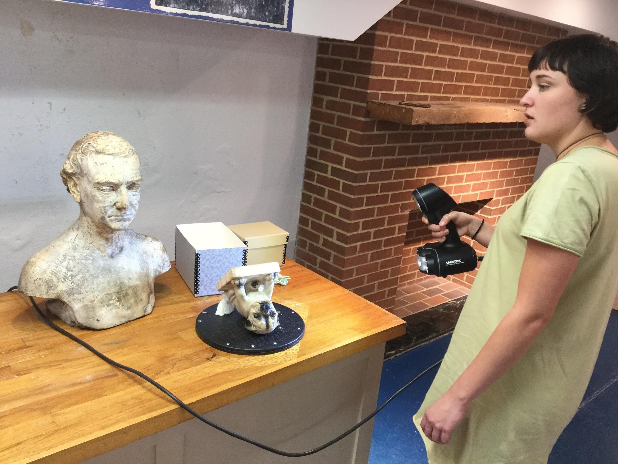 A person holds a black 3D scanner. A sculpture of a pug dog and the bust of an unknown person are seated on the table.