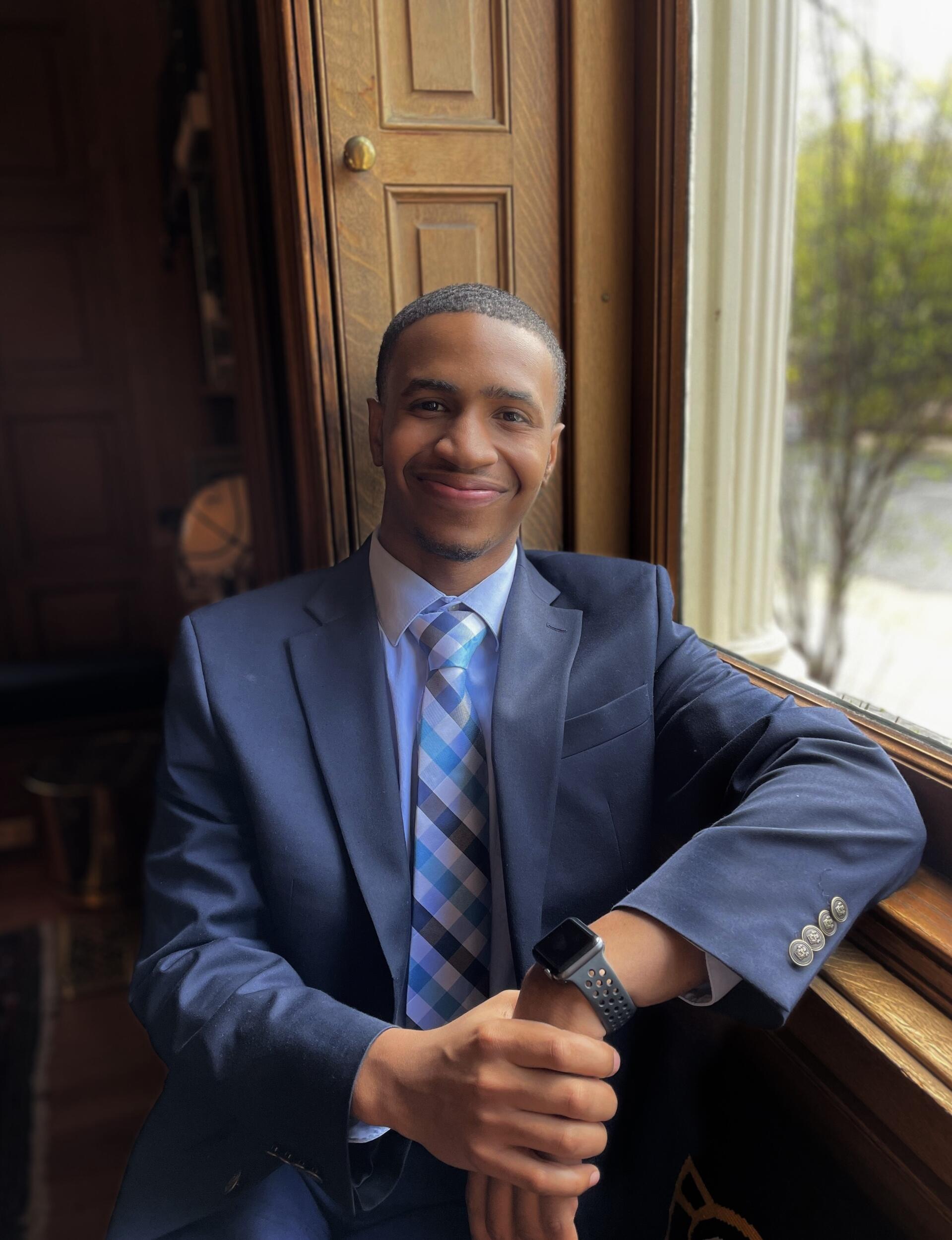 A portrait of a young man wearing a blue suit jacket and tie leaning against a window frame. 