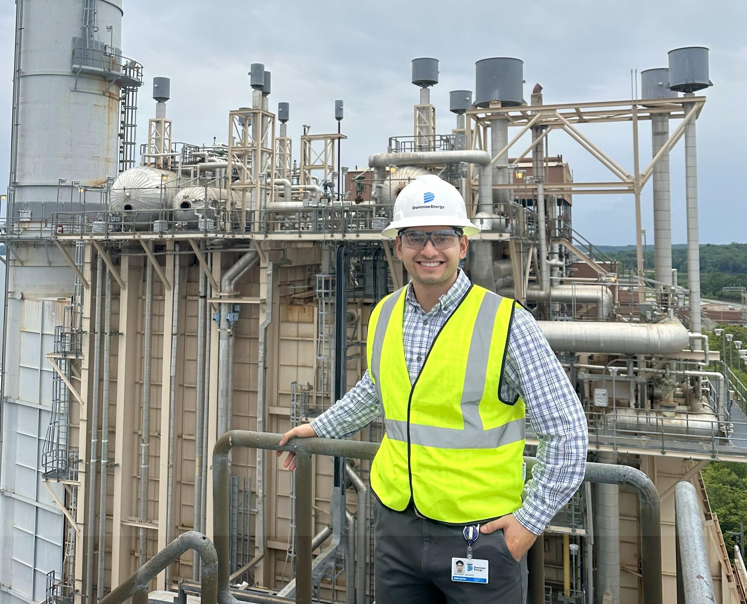 A man in a yellow safety vest and hard hat stands in front of a power plant.
