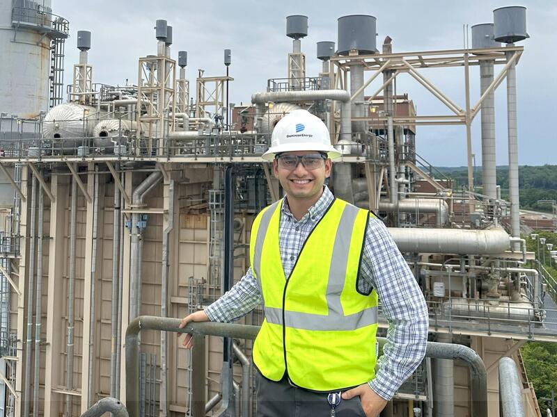 A man in a yellow safety vest and hard hat stands in front of a power plant.