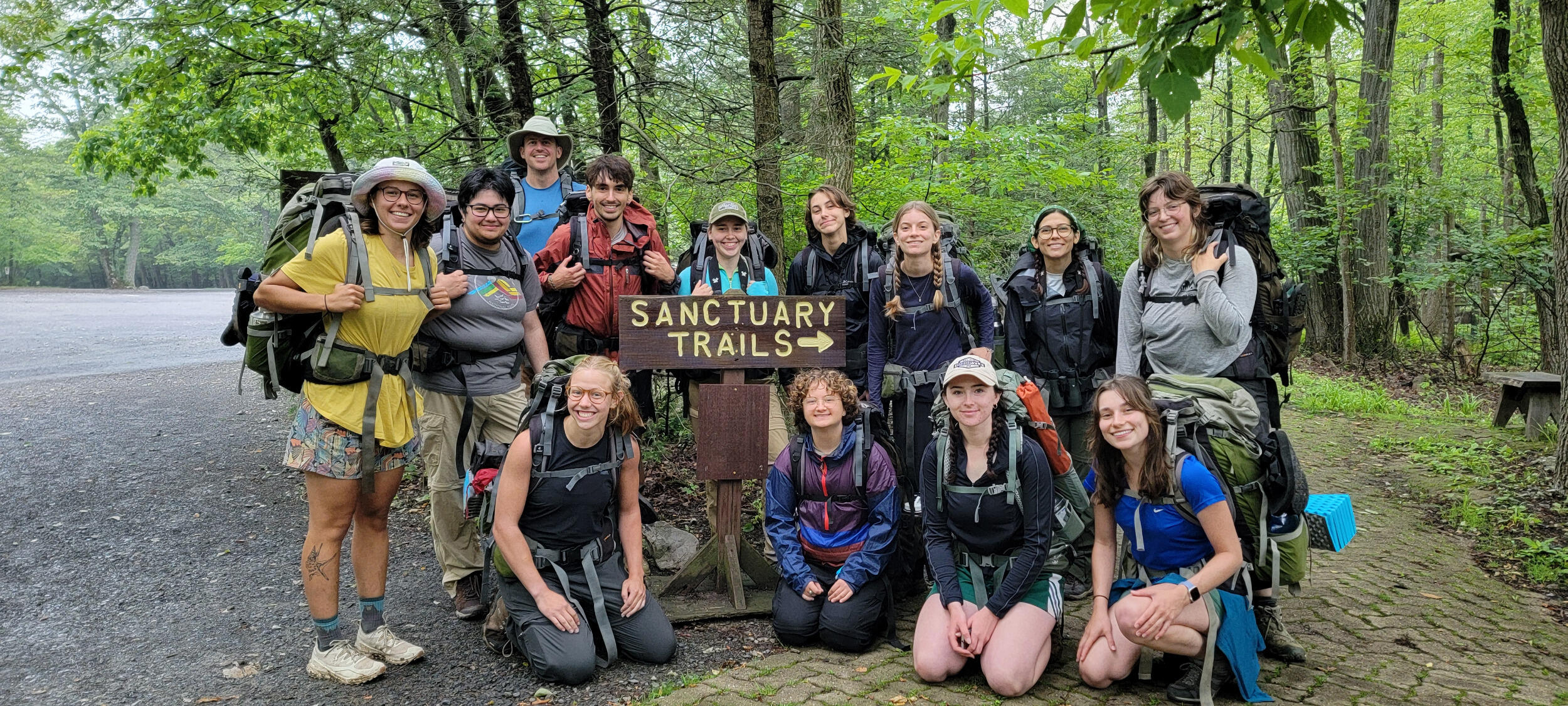 A group of people wearing hiking backpacks pose next to a trail sign and trail.