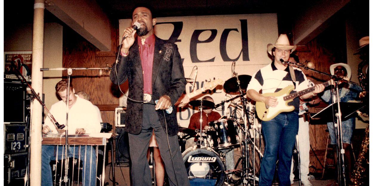 A band performing. On the left is a man on a keyboard, in the center is a man singing in the back is a man on drums, and to the right is a man on base. 