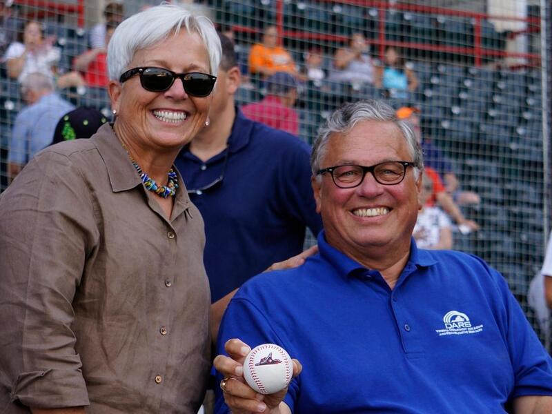 A woman wearing sunglasses smiling and standing next to a man smiling and holding a baseball. 