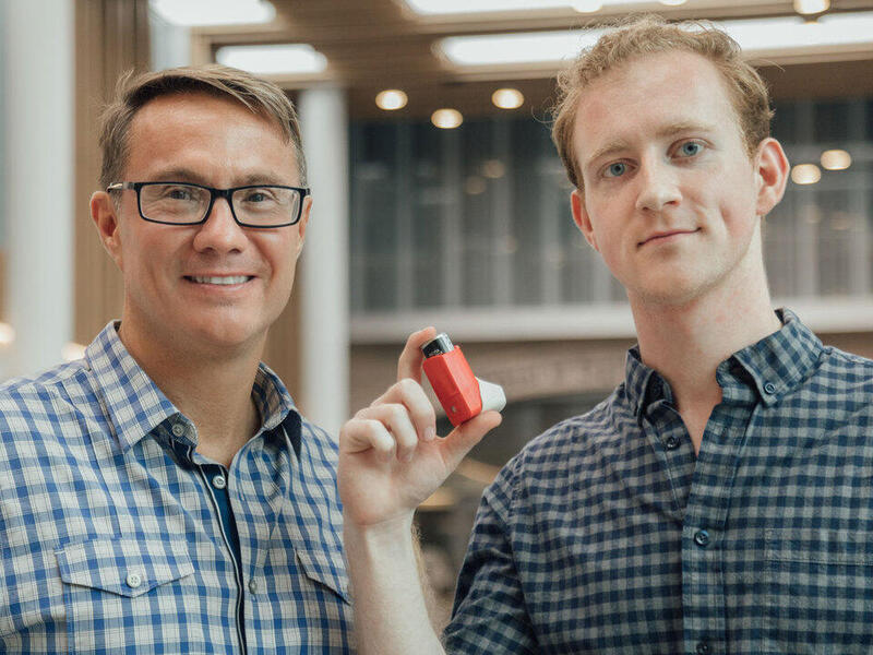 A photo of two men from the chest up. The man on the right is holding an inhaler. 
