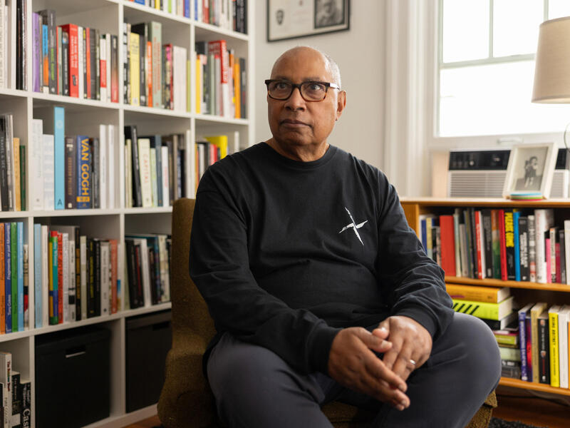 A man in glasses and a black shirt sits in a chair in front of shelves of books.