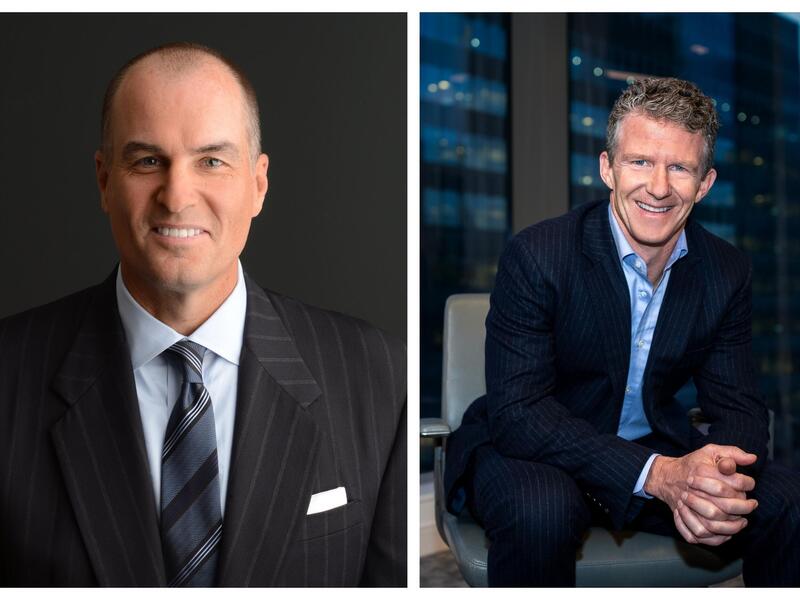 A photo of a man sitting in a chair in between two windows. The windows show a large office building behind him. 