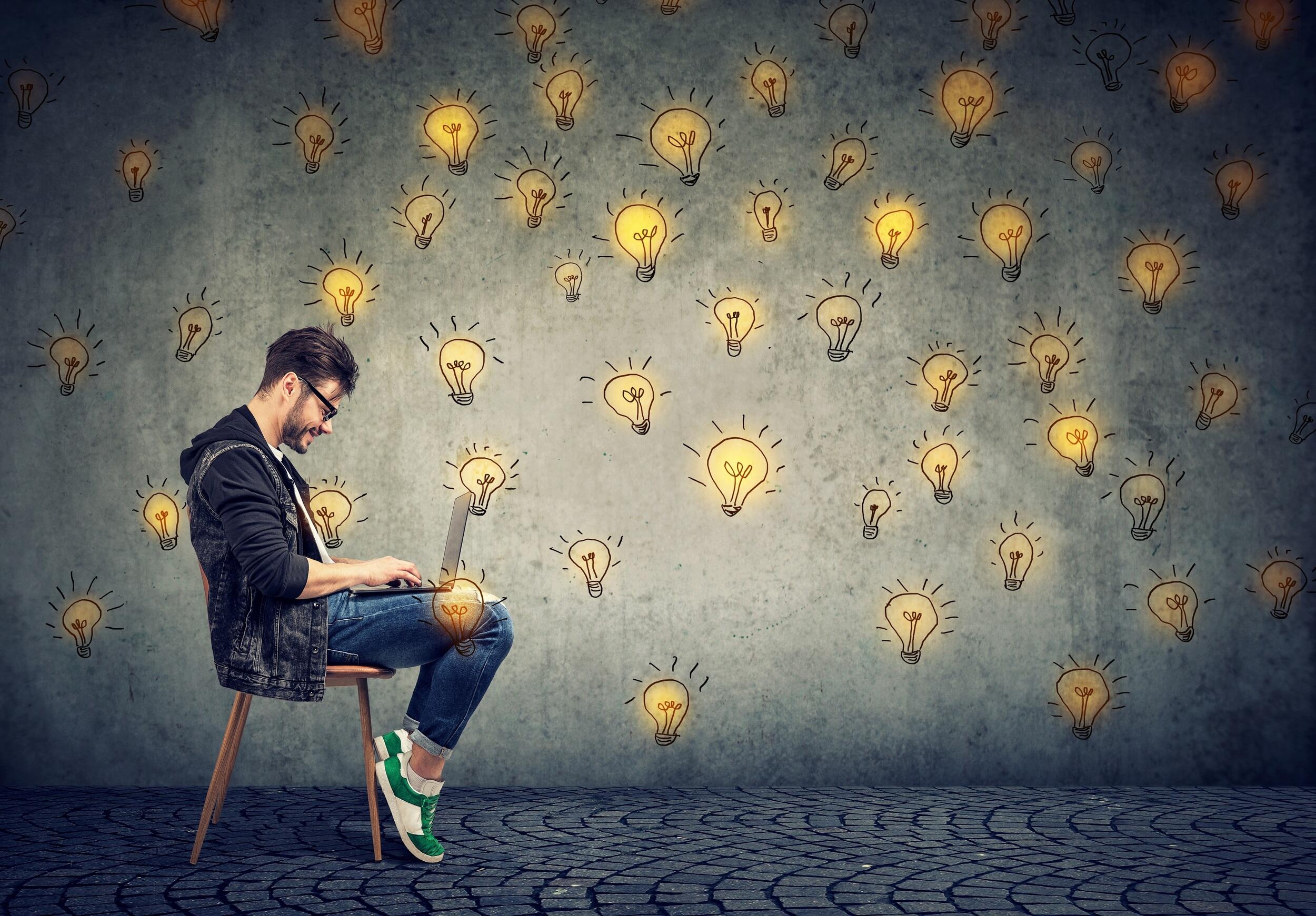 A man sitting in a chair looking at a computer with drawings of lit yellow lightbulbs displayed on the wall behind him.