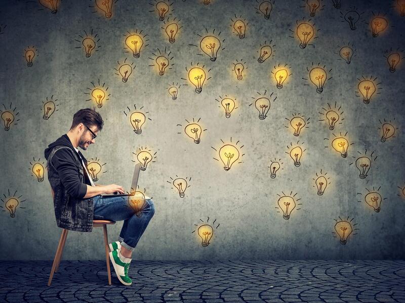 A man sitting in a chair looking at a computer with drawings of lit yellow lightbulbs displayed on the wall behind him.