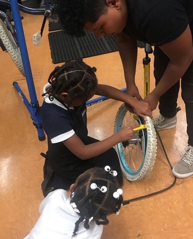 A child removes the tread of a bicycle tier with the help of a technician while another child looks on.