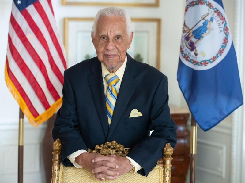 A photo of a man standing between an American flag and the Virginia state flag. 