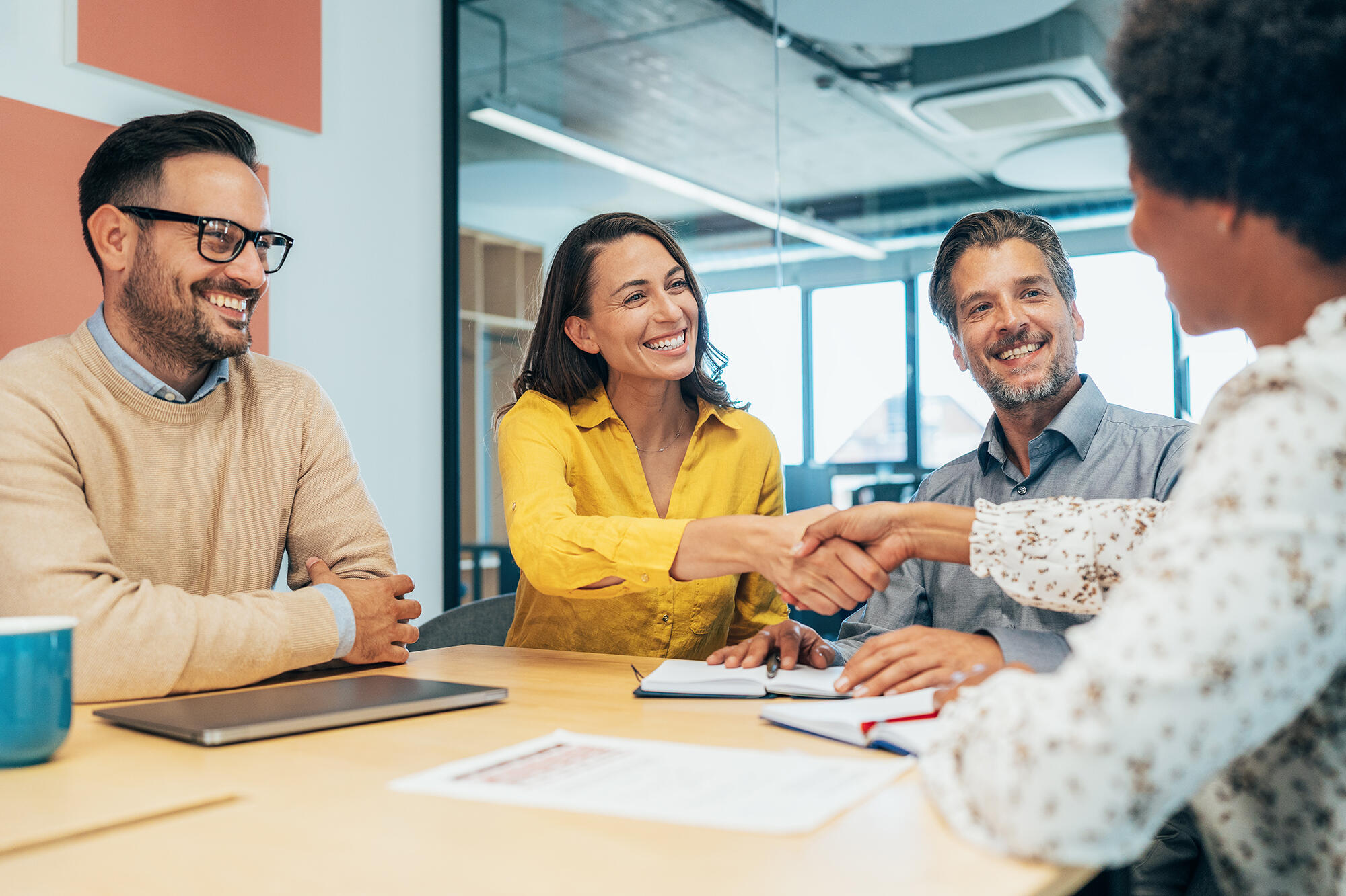 A photo of three people on one side of a table and one person sitting on the opposite side. The person in the middle on the left side and the person on the right side are shaking each others hands. 